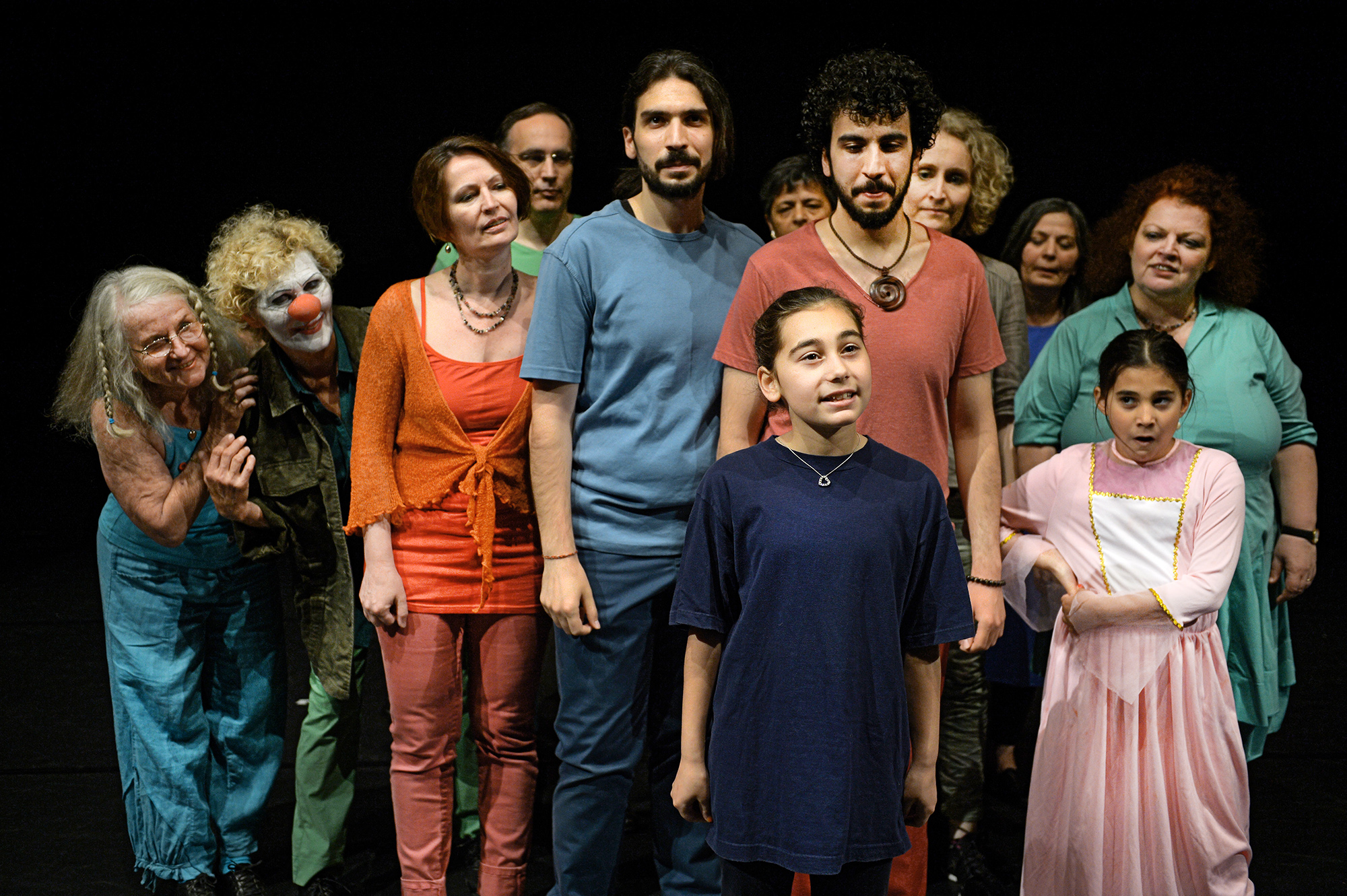 Inszenierungsfoto aus „#Widerstand“ auf einer Studiobühne mit schwarzem Holzboden. Das generationenübergreifende Ensemble versammelt sich als Gruppe um eine junge Spielerin. Die Spielerin wendet sich erzählend ans Publikum, das Ensemble richtet die Blicke auf die Spielerin. 